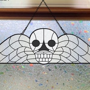 May include: A black and white stained glass pattern of a skull with wings, hanging in front of a window. The image includes the text "DIGITAL DOWNLOAD STAINED GLASS PATTERN" and "HOBBY & COMMERCIAL LICENSES INCLUDED". Two decorative glass bottles sit on the windowsill.