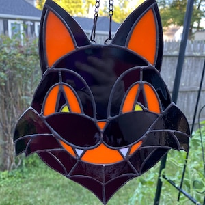 Op de afbeelding: Een zwart kattengezicht zonnevanger met oranje en gele accenten. De ogen en oren van de kat zijn oranje, met gele details. De mond heeft witte tanden en een spinnenwebontwerp. Het hangt aan een ketting, perfect voor Halloween decoratie.