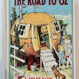 May include: A hardcover book titled The Road to Oz by L. Frank Baum. The cover illustration features a large orange pumpkin-shaped house with a chimney, a door, and windows. A pumpkin-headed character stands in the doorway. A dog and children are nearby.