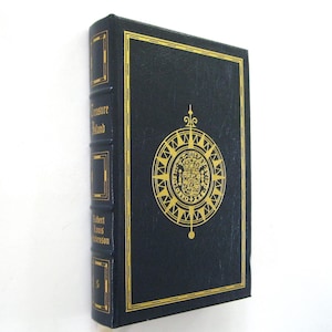 Pode incluir: Um livro de capa dura azul escuro intitulado "Treasure Island" de Robert Louis Stevenson. A capa apresenta letras douradas e um desenho de rosa dos ventos dourada. O livro est&aacute; em bom estado.