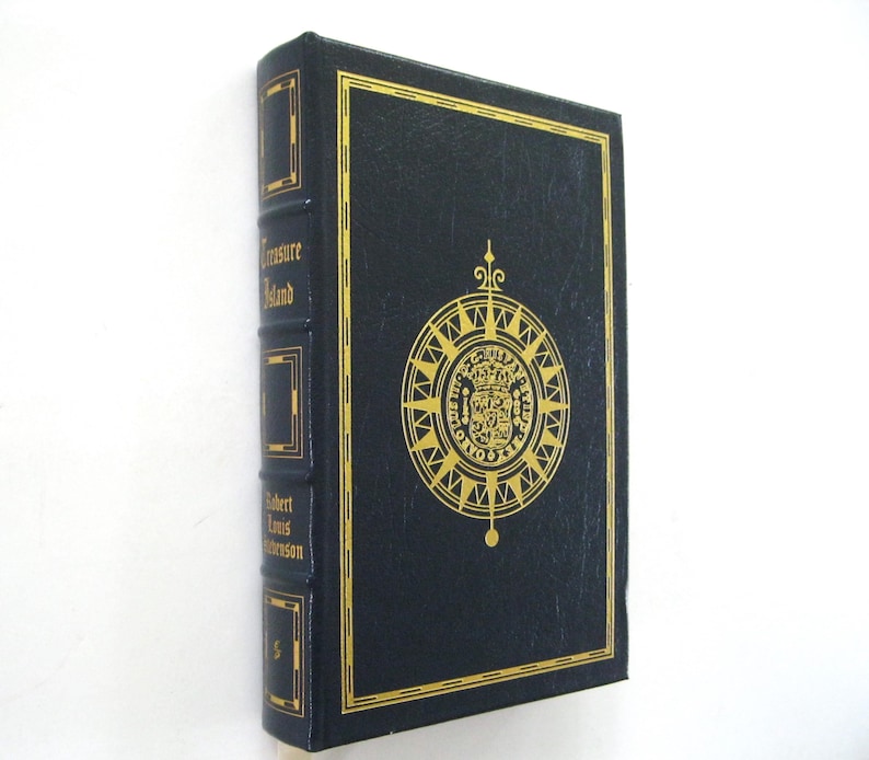 Pode incluir: Um livro de capa dura azul escuro intitulado "Treasure Island" de Robert Louis Stevenson. A capa apresenta letras douradas e um desenho de rosa dos ventos dourada. O livro est&aacute; em bom estado.