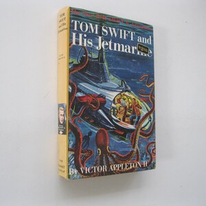 May include: Vintage hardcover book titled "Tom Swift and His Jetmarine" by Victor Appleton II. The cover features a submarine scene with an octopus and figures inside. The book has a yellow spine and a price tag of $2.00.