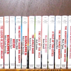 May include: A row of classic novels with white spines and colourful titles, including "Oliver Twist" and "Moby Dick." The books are displayed on a wooden shelf against a dark brown backdrop. The titles are clearly visible.