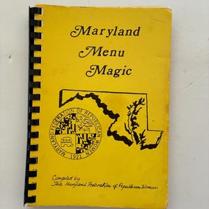 May include: A vintage cookbook titled "Maryland Menu Magic" with a yellow cover and black spiral binding. The cover features the Maryland state outline, a seal, and the text "Maryland Menu Magic". Compiled by The Maryland Federation of Republican Women.