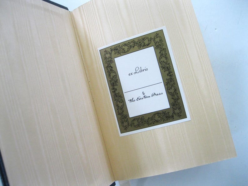 Pode incluir: Livro aberto com p&aacute;ginas claras e uma etiqueta decorativa. A etiqueta, emoldurada em dourado, diz "ex Libris" e "The Easton Press" em uma escrita elegante. As p&aacute;ginas do livro t&ecirc;m uma sutil textura de gr&atilde;o de madeira.