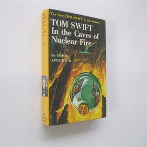 May include: A vintage hardcover book titled "TOM SWIFT In the Caves of Nuclear Fire" by Victor Appleton II. The cover features a green submarine-like vessel, a flying object, and a fiery background. The book has a yellow spine.