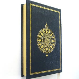 Pode incluir: Um livro de capa dura azul marinho com detalhes dourados. A capa apresenta um emblema dourado com um design de b&uacute;ssola. As p&aacute;ginas do livro t&ecirc;m bordas douradas. Uma fita branca como marcador de livro &eacute; vis&iacute;vel.