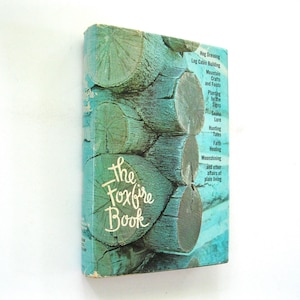 May include: The Foxfire Book, a vintage book with a teal and green cover featuring a log cabin design. The title is in white script. The spine lists topics like log cabin building, crafts, and hunting tales.