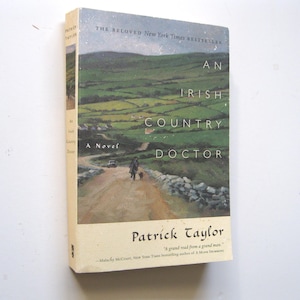 May include: A hardcover book titled "An Irish Country Doctor" by Patrick Taylor. The cover features a landscape painting of a rural road with a person and dog walking on it, and green fields in the background.