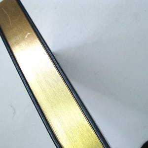 Pode incluir: Close-up da borda dourada de um livro. As bordas das p&aacute;ginas douradas s&atilde;o vis&iacute;veis, com uma capa preta. O livro est&aacute; posicionado diagonalmente contra um fundo branco, mostrando o contraste entre o ouro e o preto.