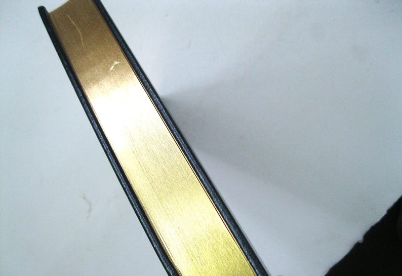 Pode incluir: Close-up da borda dourada de um livro. As bordas das p&aacute;ginas douradas s&atilde;o vis&iacute;veis, com uma capa preta. O livro est&aacute; posicionado diagonalmente contra um fundo branco, mostrando o contraste entre o ouro e o preto.