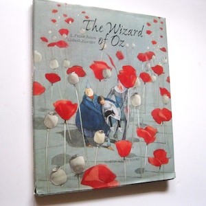 May include: A hardcover book titled "The Wizard of Oz" with a light blue cover. The cover features illustrations of red poppies and figures in blue and white clothing. The book is published by North-South Books.