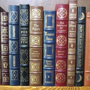 May include: A collection of antique books with various titles and decorative covers. The spines display titles like "Main Street," "Poe," and "Treasure Island." The books are in shades of brown, blue, and red, with gold lettering and embellishments.