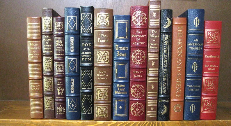 Pode incluir: Uma cole&ccedil;&atilde;o de livros antigos com capas decorativas. Os t&iacute;tulos incluem "Main Street", "Poe" e "Treasure Island". As lombadas s&atilde;o castanhas, azuis e vermelhas, com letras douradas e padr&otilde;es. Os livros est&atilde;o dispostos numa prateleira.