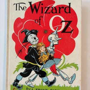 May include: A hardcover book titled The Wizard of Oz by L. Frank Baum with illustrations by W.W. Denslow. The cover features a red poppy background with the Scarecrow, Dorothy holding a dog, and the Tin Woodman walking across a green field.