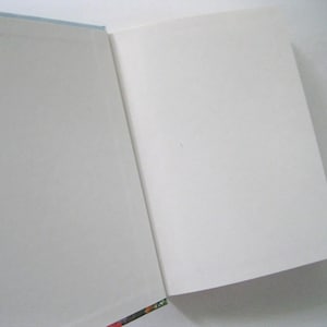 May include: An open book with blank white pages. The book's spine is visible on the left side, with a colorful floral pattern. The pages are clean and ready for writing or reading. The book's edges are a light blue.
