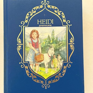 May include: A hardcover book titled "Heidi" by Johanna Spyri. The cover is a deep blue with a gold decorative frame. The illustration depicts a young girl with a goat and a basket, set against a mountain backdrop.
