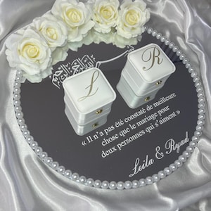 May include: A mirrored tray with a pearl border, holding two white ring boxes with gold lettering. The tray is decorated with white roses and text in French. The boxes are labeled "L" and "R".