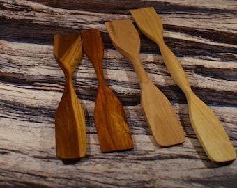 May include: Four wooden spatulas with wide, rectangular heads and long, slender handles. The spatulas are made of light and dark wood, with visible grain patterns. They are arranged on a textured, wooden surface.