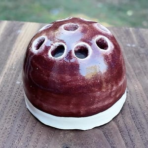 Könnte beinhalten: Ein bordeauxfarbener Keramik-Blumenfrosch mit mehreren Löchern oben, der auf einer Holzoberfläche sitzt. Der Blumenfrosch hat einen weißen Sockel und ein glänzendes Finish, das für die Anordnung von Blumen bestimmt ist.