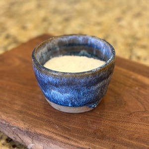 May include: A handmade ceramic bowl with a blue and brown glaze, filled with a white powder. The bowl sits on a wooden cutting board with a natural edge. The bowl's interior is a light color, contrasting with the darker exterior.