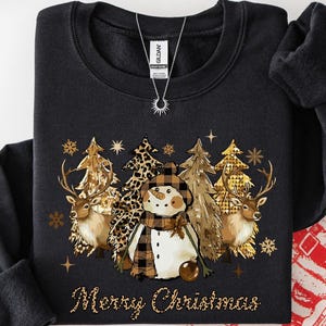May include: Black crew neck sweatshirt with a festive Christmas design. The graphic features a snowman, deer, and trees with gold accents, and the words "Merry Christmas." A necklace with a sun pendant is visible. A red and white Christmas bag is in the background.