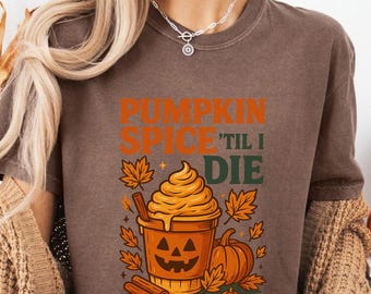 May include: A brown t-shirt with the text "PUMPKIN SPICE 'TIL I DIE" and a graphic of a pumpkin spice latte in a jack-o'-lantern cup. The shirt is paired with black pants and a tan sweater. Pumpkins and fall decor are in the background.