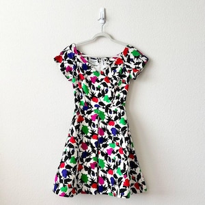 Puede incluir: Un vestido con estampado floral con escote en V y mangas cortas. El vestido presenta un fondo blanco con un estampado de flores coloridas en rojo, rosa, verde y azul, y hojas negras. El vestido está colgado en una percha blanca.