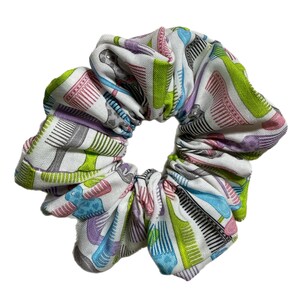 May include: A white scrunchie with a repeating pattern of colorful combs in shades of green, pink, purple, blue, and black. The hair accessory is made of fabric and is designed to hold hair in a ponytail or bun.