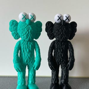 May include: Two toy figures, one teal and one black, are displayed. Each has a textured, fur-like appearance, with large, round white eyes featuring an 'X' design. The teal figure has green 'X' eyes, while the black figure has black 'X' eyes.