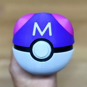 May include: A spherical object featuring a purple top section with pink accents and a white bottom section. A black horizontal band separates the two halves, featuring a central white circular button. The letter M is printed in white on the top.