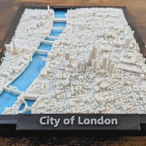 Pode incluir: Um modelo 3D da cidade de Londres, apresentando uma paisagem urbana branca detalhada com um rio azul. O modelo está em uma moldura preta, com as palavras "City of London" exibidas em uma pequena placa.