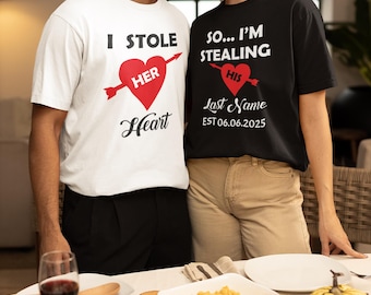Puede incluir: Camisetas a juego, blancas y negras, con texto romántico. La camiseta blanca dice "I Stole Her Heart" con un corazón rojo atravesado por una flecha. La camiseta negra dice "So... I'm Stealing His Last Name" con un corazón rojo y una fecha.