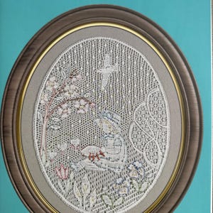 May include: An oval framed piece of lace art featuring a detailed scene of a person in a hat, surrounded by flowers and a bird. The frame is brown with a gold inner edge, set against a teal background. The text "Little Lacemakers" is at the top.