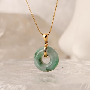 May include: A gold chain necklace featuring a circular green jade pendant. The pendant is suspended from a gold bail that connects to the thin, delicate gold chain. The jade stone displays natural green and white mottled color variations.