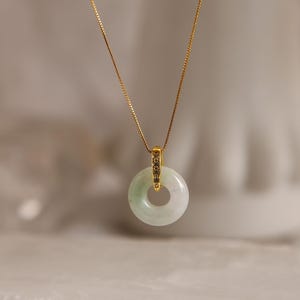 May include: A delicate gold chain necklace featuring a small, circular, pale green jade pendant. The pendant is suspended from a gold bail that connects to the thin, metallic chain, creating a simple and refined piece of jewelry for the neck.