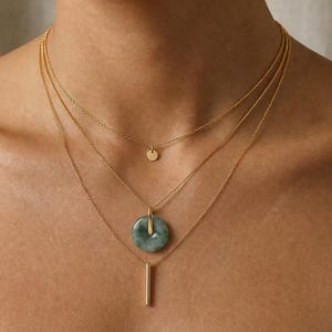 May include: A layered gold necklace set featuring three delicate chains. The top chain has a small circular pendant, the middle chain has a green jade pendant, and the bottom chain has a vertical bar pendant. The necklaces are displayed on a person's neck.