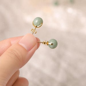 May include: A pair of gold stud earrings, each with a round, pale green jade stone. The earrings have a delicate, minimalist design with a gold setting that cradles the jade sphere. The earrings are held up by a hand.