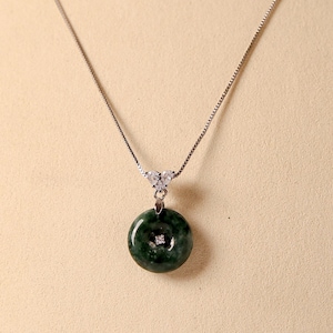Puede incluir: Collar de plata con un colgante de jade verde oscuro y un pequeño diamante. El colgante circular tiene un agujero en el centro. La delicada cadena cuelga de una fianza en forma de corazón. Una joya elegante.