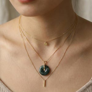 May include: A layered necklace set featuring three gold chains. The top chain has a small, round pendant. The middle chain holds a dark green jade pendant. The bottom chain has a vertical bar pendant.