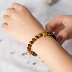 May include: A beaded bracelet featuring tiger's eye stones and a geometric yellow stone. The bracelet is worn on a wrist, showcasing the natural colors and textures of the gemstones. The bracelet is a piece of jewelry.