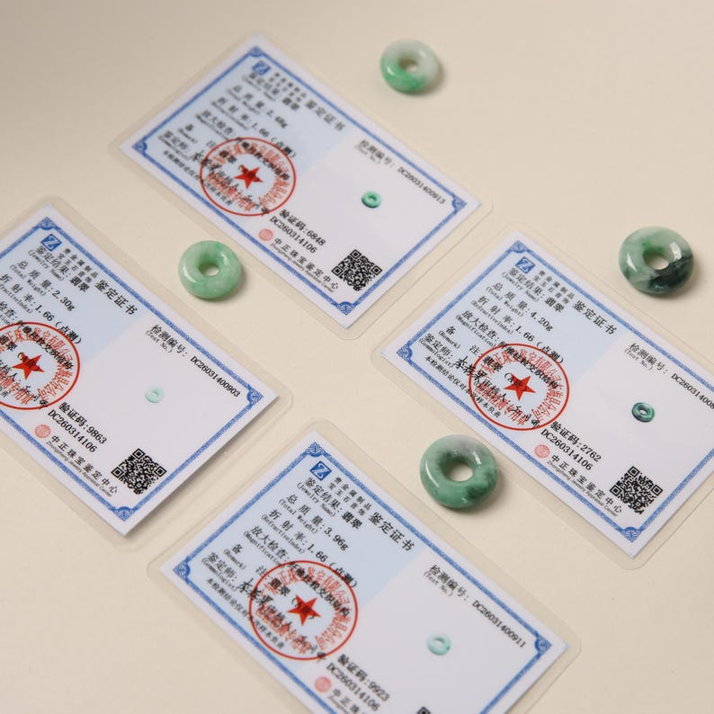 K&ouml;nnte beinhalten: Vier Jade-Donut-Anh&auml;nger mit Zertifikaten. Der Jade ist hellgr&uuml;n mit dunkleren gr&uuml;nen Maserungen. Die Zertifikate sind wei&szlig; mit chinesischem Text und roten Siegeln. Der Hintergrund ist hellbeige.