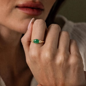 Pode incluir: Um anel de ouro com uma pedra preciosa verde de formato oval ao centro. A banda apresenta um design texturizado e canelado em ambos os lados da pedra, acentuado com pequenas pedras claras e brilhantes incrustadas no metal.