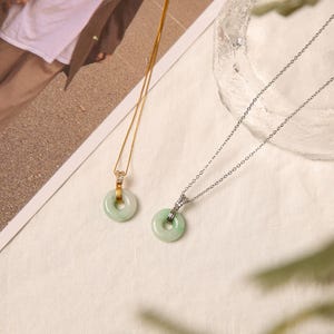 K&ouml;nnte beinhalten: Zwei Jade-Donut-Anh&auml;nger-Halsketten. Eine hat eine goldene Kette und die andere eine silberne Kette. Beide Anh&auml;nger sind hellgr&uuml;n mit einem kreisf&ouml;rmigen Design. Die Halsketten werden auf einer wei&szlig;en Oberfl&auml;che pr&auml;sentiert.