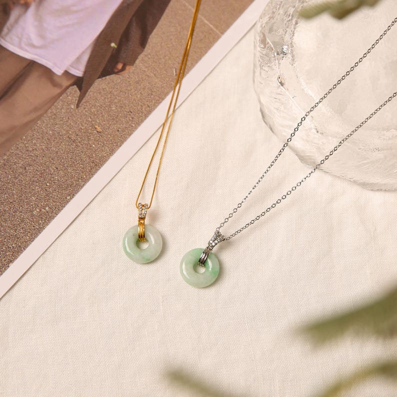 K&ouml;nnte beinhalten: Zwei Jade-Donut-Anh&auml;nger-Halsketten. Eine hat eine goldene Kette und die andere eine silberne Kette. Beide Anh&auml;nger sind hellgr&uuml;n mit einem kreisf&ouml;rmigen Design. Die Halsketten werden auf einer wei&szlig;en Oberfl&auml;che pr&auml;sentiert.