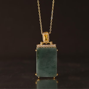 May include: A gold-colored chain necklace with a rectangular, dark green pendant. The pendant is framed with gold and small, clear stones. The top of the pendant has a gold-colored accent with a yellow stone.