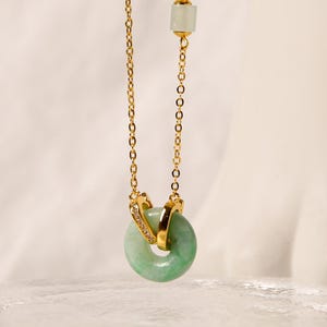 May include: A gold-toned necklace featuring a light green jade donut pendant. The pendant is encircled by a gold band with a small section of clear crystals. The necklace chain is also gold, with a small jade bead at the top.