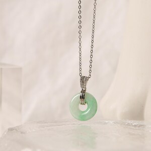 K&ouml;nnte beinhalten: Eine silberne Halskette mit einem hellgr&uuml;nen Jade-Donut-Anh&auml;nger. Der Anh&auml;nger h&auml;ngt an einer feinen Kette und hat eine polierte Oberfl&auml;che. Die Halskette wird vor einem weichen, neutralen Hintergrund pr&auml;sentiert. Der Jade-Anh&auml;nger hat einen Durchmesser von ca. 2,5 cm.