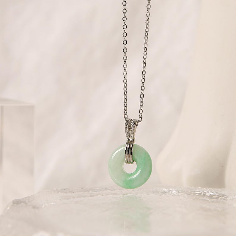 K&ouml;nnte beinhalten: Eine silberne Halskette mit einem hellgr&uuml;nen Jade-Donut-Anh&auml;nger. Der Anh&auml;nger h&auml;ngt an einer feinen Kette und hat eine polierte Oberfl&auml;che. Die Halskette wird vor einem weichen, neutralen Hintergrund pr&auml;sentiert. Der Jade-Anh&auml;nger hat einen Durchmesser von ca. 2,5 cm.