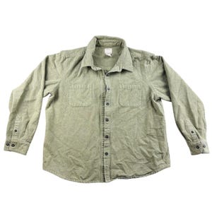 May include: A long-sleeved shirt in a muted olive green hue. It has a button-down front, two chest pockets, and buttoned cuffs. The shirt features a collared neckline and is made from a soft, textured fabric.